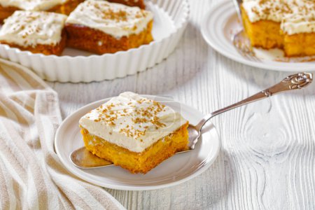 pumpkin cake with cream frosting in baking pan and one slice on a white plate, on white wooden table horizontal view from above
