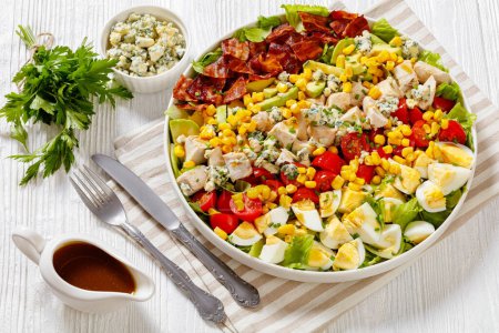 American garden salad of chopped romaine lettuce, tomato, crisp bacon, chicken breast, hard-boiled eggs, avocado, blue cheese, and red-wine vinaigrette on plate on white wooden table