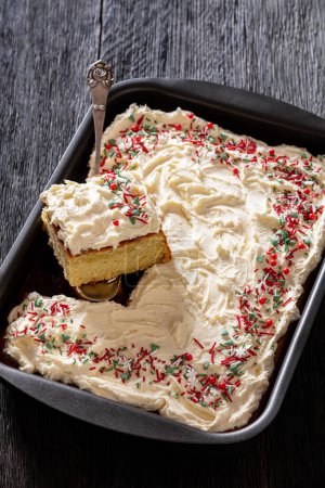 christmas vanilla sheet cake with whipped buttercream frosting garnished with sprinkles in baking dish with cake shovel on dark wooden table, vertical view