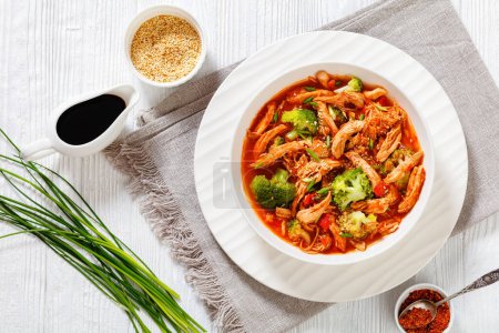 slow-cooked shredded chicken breast stew with tomato sauce, red pepper, onion and broccoli in white bowl on wood table, horizontal view from above, flat lay