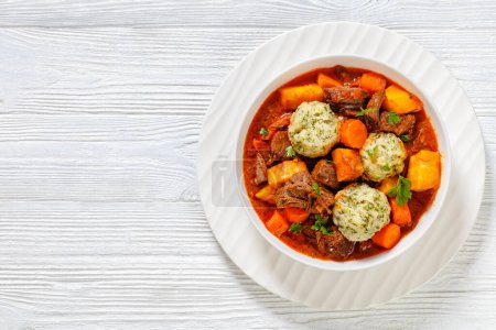 Beef Stew with Dumplings and vegetables in rich tomato and stock based gravy in white bowl, british cuisine, horizontal view from above, flat lay, free space, close-up