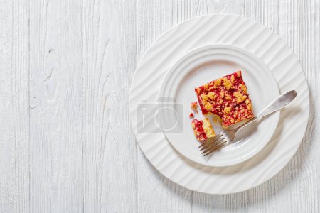 Himbeer-Crumble-Riegel auf weißem Teller mit Dessertgabel auf weißem Holztisch, Horizontalblick von oben, flache Lage, freier Raum