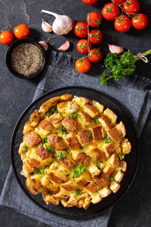 cheesy garlic bread, pull apart whole round bread on black platter on concrete table with melted butter, tomatoes, and spices at background, vertical view from above