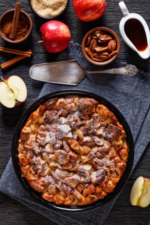Cinnamon Apple French Toast Bake topped with maple syrup and toasted pecans in baking dish on dark wooden table with ingredients, vertical view from above