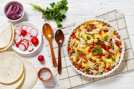 shredded chicken breast, pinto beans, tomato and mozzarella in white round baking dish on white wood table with wooden spoons, flatbreads, red radish, horizontal view from above, flat lay