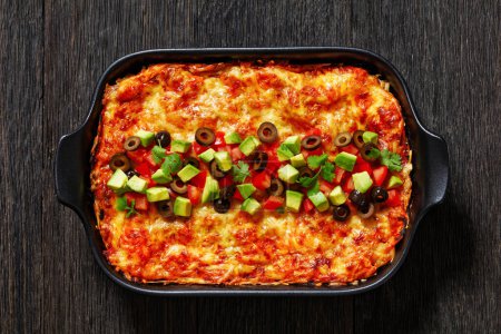 Healthy chicken and bean gluten free lasagna topped with pitted olives, avocado, tomatoes and coriander in black baking dish on dark wood table, horizontal view from above, flat lay, close-up