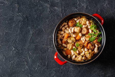 french cassoulet of chicken breast meat, sausages, white beans and vegetables in red pot on grey concrete table, horizontal view from above, flat lay, free space