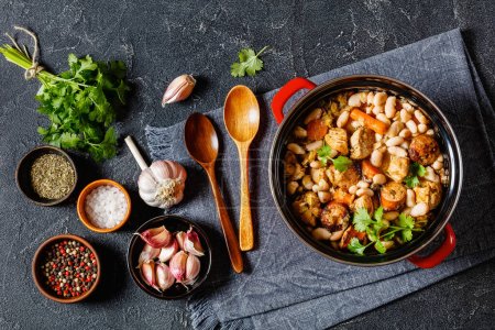 french cassoulet of chicken breast meat, sausages, white beans and vegetables in red pot on grey concrete table with ingredients and wood spoons, horizontal view from above, flat lay