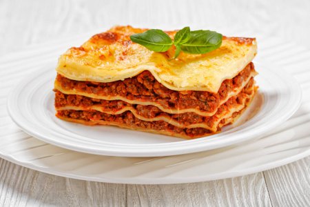 portion of lasagne al forno, italian beef lasagna layered with ground beef, marinara sauce, pasta noodles and ricotta cheese on white plate on white wood table, close-up
