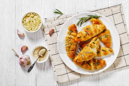 savory crescent rolls with melted cheese filling, sprinkled with fresh rosemary leaves on white plate on white wooden table with garlic butter, horizontal view from above, flat lay