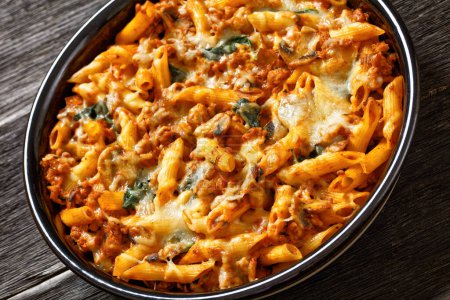 close-up of Ground Chicken Pasta Bake with onion, mushrooms, spinach, tomato sauce and mozzarella cheese in baking dish on dark wood table