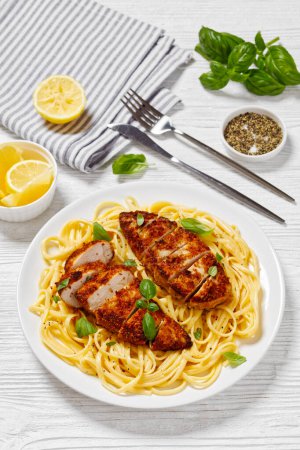 Chicken Scallopini of crusted chicken breast over lemon butter pasta on white plate on white wooden table with cutlery, vertical view, close-up