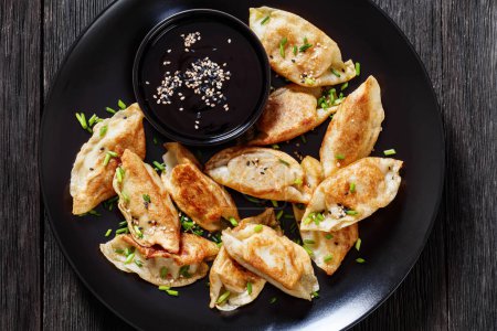 fried gyoza, wonton wrappers stuffed with pork and cabbage sprinkled with green onions on black plate with soy sauce on dark wooden table, flat lay, close-up