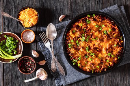 Cajun Beef Cornbread Casserole topped with melted red cheddar cheese and scalions in baking dish on dark wood table with ingredients and seasoning, horizontal view, flat lay