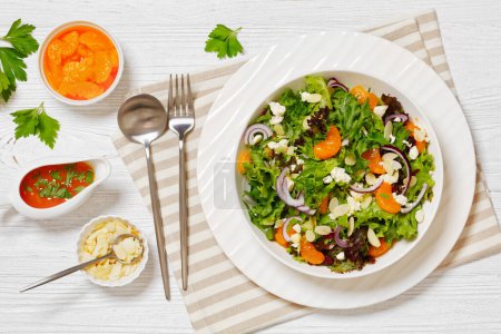 Mandarin Orange Salad of lettuce mix, toasted almonds, dried cranberries, red onion and feta cheese in white bowl on white textured wooden table, horizontal view from above, flat lay