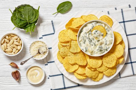 artichoke spinach almond creamy dip with potato chips on white platter on white wooden table with ingredients, horizontal view from above, flat lay
