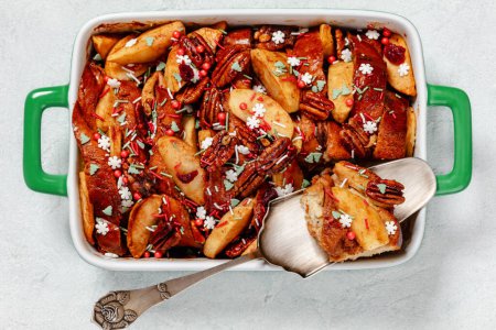 Baked Apple French Toast topped with pecan nuts and christmas sugar sprinkles in baking dish on white concrete table, horizontal view from above, flat lay, close-up