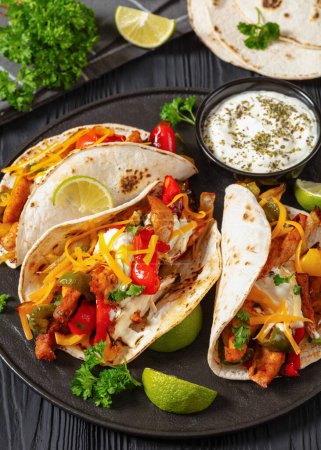 baked tex-mex chicken fajitas with mixed sweet pepper, onion, sour cream, shredded cheese and white corn tortillas on black plate on black wooden table, vertical view, close-up