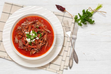 birria de res, mexican beef stew in hot red pepper sauce with raw onion and chopped cilantro in white bowl on white wooden table with forks, horizontal view from above, flat lay, free space