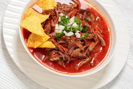 birria de res, mexican beef stew in hot red pepper sauce with raw onion and chopped cilantro, taco chips in white bowl, close-up