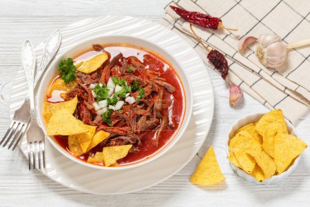 birria de res, mexican beef stew in hot red pepper sauce with raw onion and chopped cilantro, taco chips in white bowl, landscape view