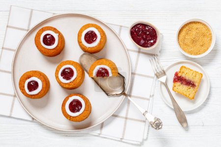 runebergin torttu, runeberg cakes, a finnish mini cake flavored with almond, raspberry jam and rum on plate on white wooden table with ingredients, horizontal view, flat lay
