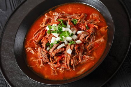 close-up of birria de res, mexican beef stew in hot red pepper sauce with raw onion and chopped cilantro in black bowl on black wooden table