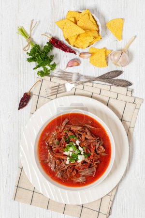 birria de res, mexican beef stew in hot red pepper sauce with raw onion and chopped cilantro in white bowl on white wooden table with taco chips and ingredients, vertical view from above