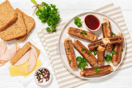 ham and cheese french whole wheat toast roll ups served with sweet chili sauce on plate on white wooden table with ingredients, horizontal view from above, flat lay