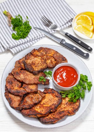 fried juicy pork steaks with ketchup and parsley on plate on white wooden table with cutlery, napkin and lemon slices, vertical view from above