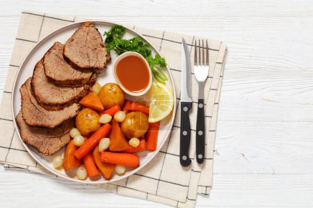 roast beef slices with dipping sauce and winter vegetables, potatoes, carrots, pearl onions, and butternut squash on platter on white wooden table with cutlery, top view, flat lay, free space