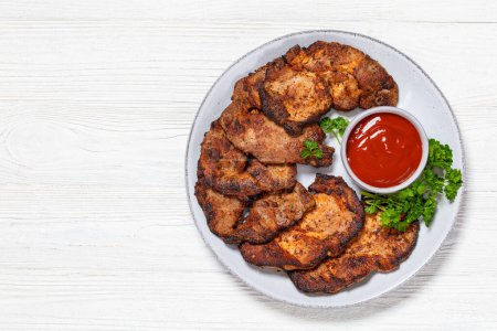 fried juicy pork steaks with ketchup and parsley on plate on white wooden table, horizontal view from above, free space, flat lay