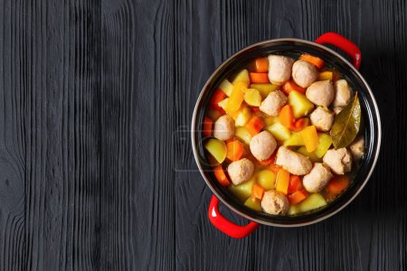 siskonmakkarakeitto, finnish sausage soup with rutabaga, potato, onion, carrot, parsnip in red pot on black wooden table, horizontal view from above, flat lay, free space