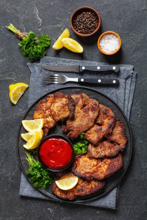 fried juicy pork steaks with ketchup, lemon slices and parsley on plate on concrete table with cutlery, salt and peppercorns, vertical view from above