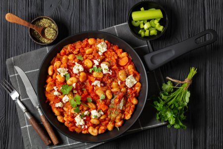 gigantes plaki, greek giant baked beans in a chunky tomato sauce sprinkled with feta cheese and fresh cilantro on skillet on black wooden table with rustic fork and knife, horizontal view, flat lay