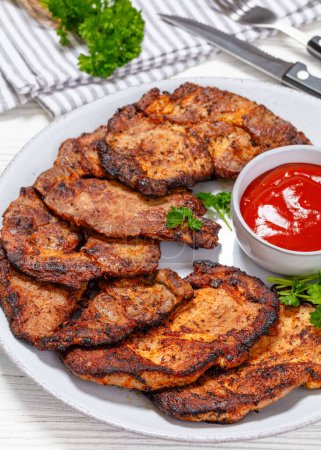 fried juicy pork steaks with ketchup and parsley on plate on white wooden table with cutlery, napkin at background, vertical view from above, close-up