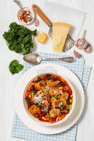 ribollita, tuscan vegetarian soup of white cannellini beans, kale, onions, carrots, celery, bread, tomatoes in white bowl on white wooden table with ingredients, vertical view from above