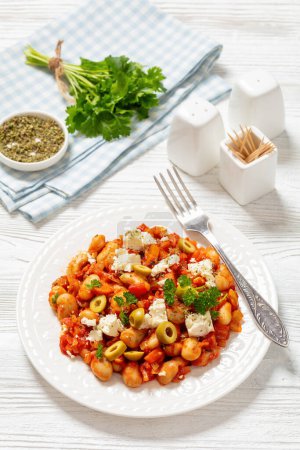 gigantes plaki, greek giant baked beans in a chunky tomato sauce sprinkled with feta cheese, olives, and fresh parsley on plate with fork on white wooden table, vertical view from above