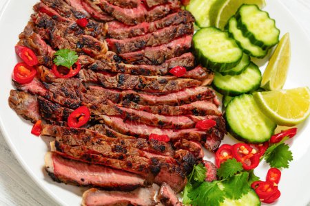 crying tiger, grilled and thinly sliced ribeye steak with cucumber and lime on plate on white wooden table with spicy dipping sauce, thai cuisine, close-up