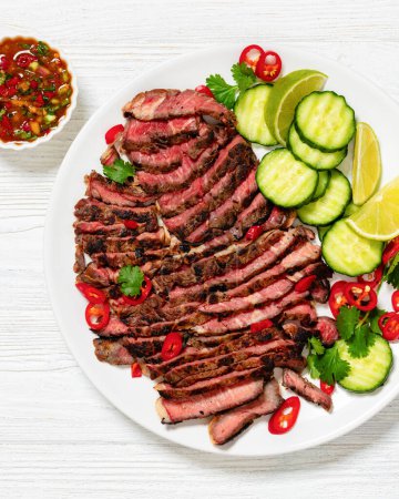 crying tiger, grilled and thinly sliced ribeye steak with cucumber and lime on plate on white wooden table with spicy dipping sauce, thai cuisine, flat lay, close-up