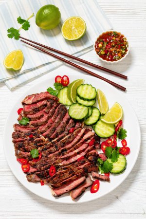 crying tiger, grilled and thinly sliced ribeye steak with cucumber and lime on plate on white wooden table with spicy dipping sauce, thai cuisine, vertical view from above