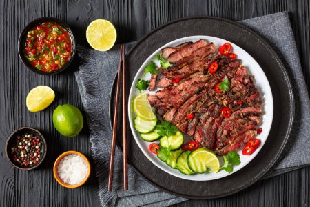 crying tiger, grilled and thinly sliced ribeye steak with cucumber and lime on plate on black wooden table with spicy dipping sauce, thai cuisine, horizontal view from above, flat lay