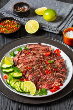 crying tiger, grilled and thinly sliced ribeye steak with cucumber and lime on plate on black wooden table with spicy dipping sauce, thai cuisine, vertical view from above, close-up