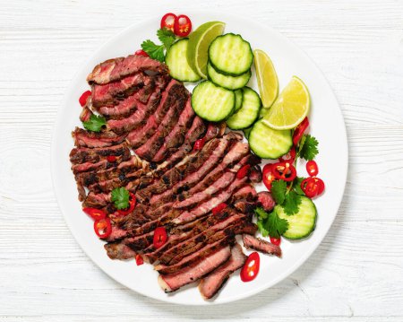 crying tiger, grilled and thinly sliced ribeye steak with cucumber and lime on plate on white wooden table with spicy dipping sauce, thai cuisine, horizontal view from above, flat lay