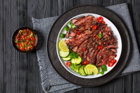 crying tiger, grilled and thinly sliced ribeye steak with cucumber and lime on plate on black wooden table with spicy dipping sauce, thai cuisine, flat lay