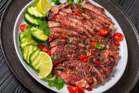 crying tiger, grilled and thinly sliced ribeye steak with cucumber and lime on plate on black wooden table, thai cuisine, close-up