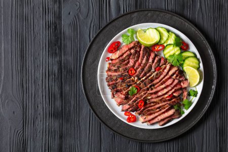 crying tiger, grilled and thinly sliced ribeye steak with cucumber and lime on plate on black wooden table with spicy dipping sauce, thai cuisine, horizontal view from above, flat lay, free space