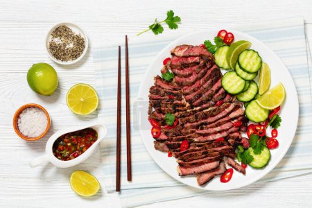 crying tiger, grilled and thinly sliced ribeye steak with cucumber and lime on plate on white wooden table with spicy dipping sauce, thai cuisine, flat lay
