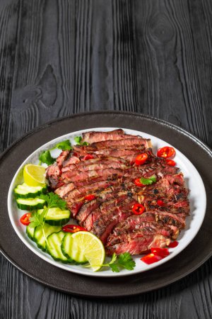 crying tiger, grilled and thinly sliced ribeye steak with cucumber and lime on plate on black wooden table, thai cuisine, vertical view, free space