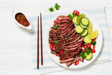 crying tiger, grilled and thinly sliced ribeye steak with cucumber and lime on plate on white wooden table with spicy dipping sauce, thai cuisine, free space, flat lay